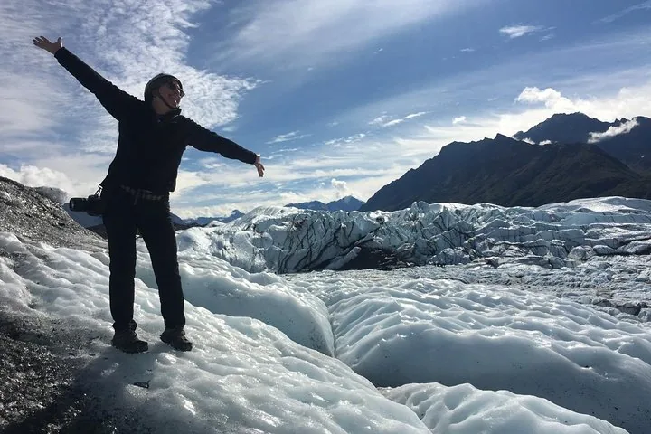 Eine Frau steht mit ausgestreckten Armen auf einem Gletscher, mit Bergen im Hintergrund.