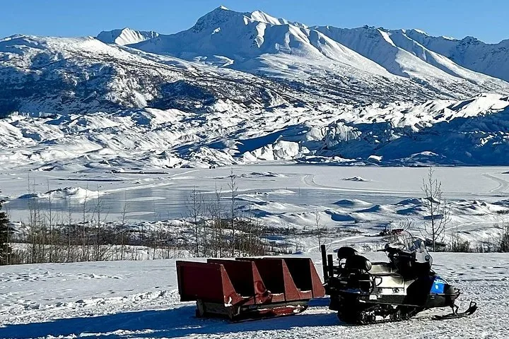 Motorschlitten und Schlitten auf Schnee geparkt, mit einem Gletscher und Bergen im Hintergrund.